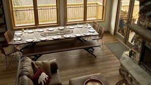 A spacious living room with a large dining table set for six, a fireplace, and a balcony with a view of mountains. - Great Family Vacay! Luxury 6 BR 300 ft to Cupp Run Hot Tub In&Outdoor Fireplace (Snowshoe)