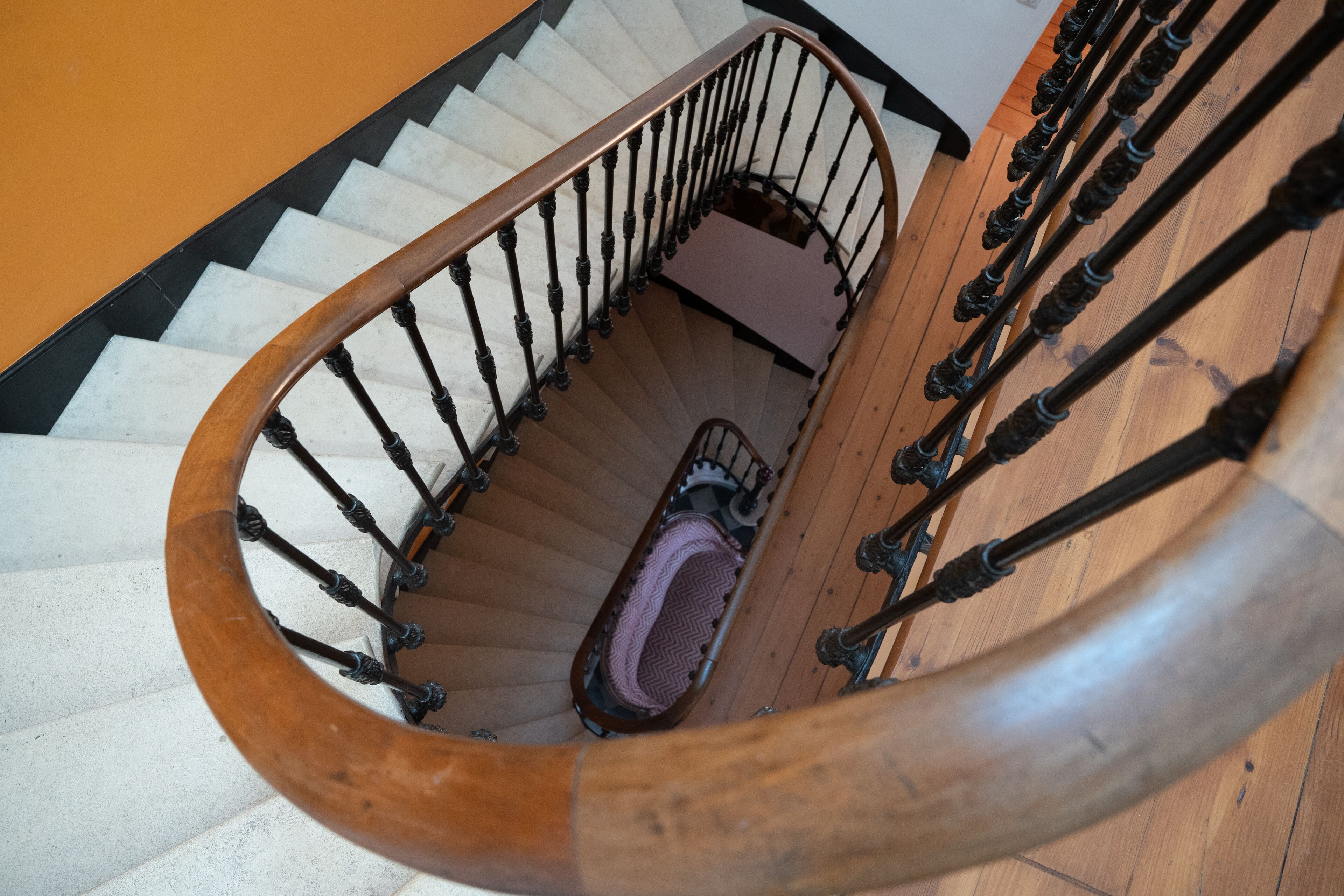 A spiral staircase with a wooden handrail and black balusters, leading to a landing with a patterned rug.