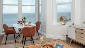 Ein Essbereich mit großem Fenster zum Meer, einem runden Tisch mit Stühlen und einer Holzkommode mit Blumen.