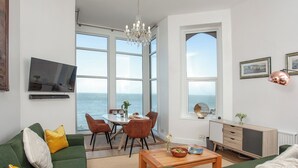 Ein modernes Wohnzimmer mit großem Meerblick, einem Essbereich mit Tisch und Stühlen sowie einer gemütlichen Sitzecke mit grünen Sofas und gelben Kissen.