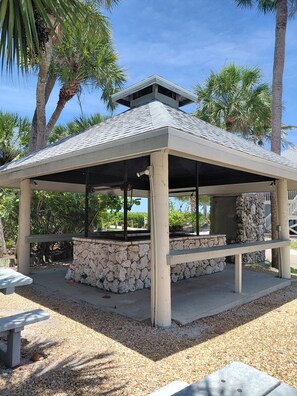 A covered outdoor area with a stone base, benches, and palm trees in the background.