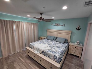 Un dormitorio con cama, mesitas de noche, ventilador de techo y ventana con cortinas.