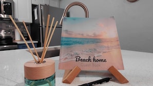 A kitchen with white cabinets, a stainless steel refrigerator, a wooden rack, a decorative wall piece, a glass container with reed diffusers, and a framed picture titled 'Beach home Guest Book'. - Luxury Condo on Little Gasparilla Island-Family Friendly, ferry included (Placida)