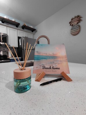 A kitchen with white cabinets, a stainless steel refrigerator, a wooden rack, a decorative wall piece, a glass container with reed diffusers, and a framed picture titled 'Beach home Guest Book'.