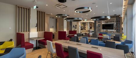 A modern dining area with a long wooden table, red and blue chairs, and black circular ceiling lights.
