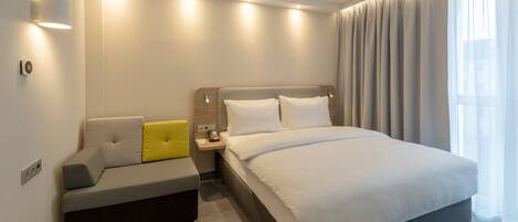 A modern hotel room with a bed, a sofa, a bedside table, and a window with curtains.
