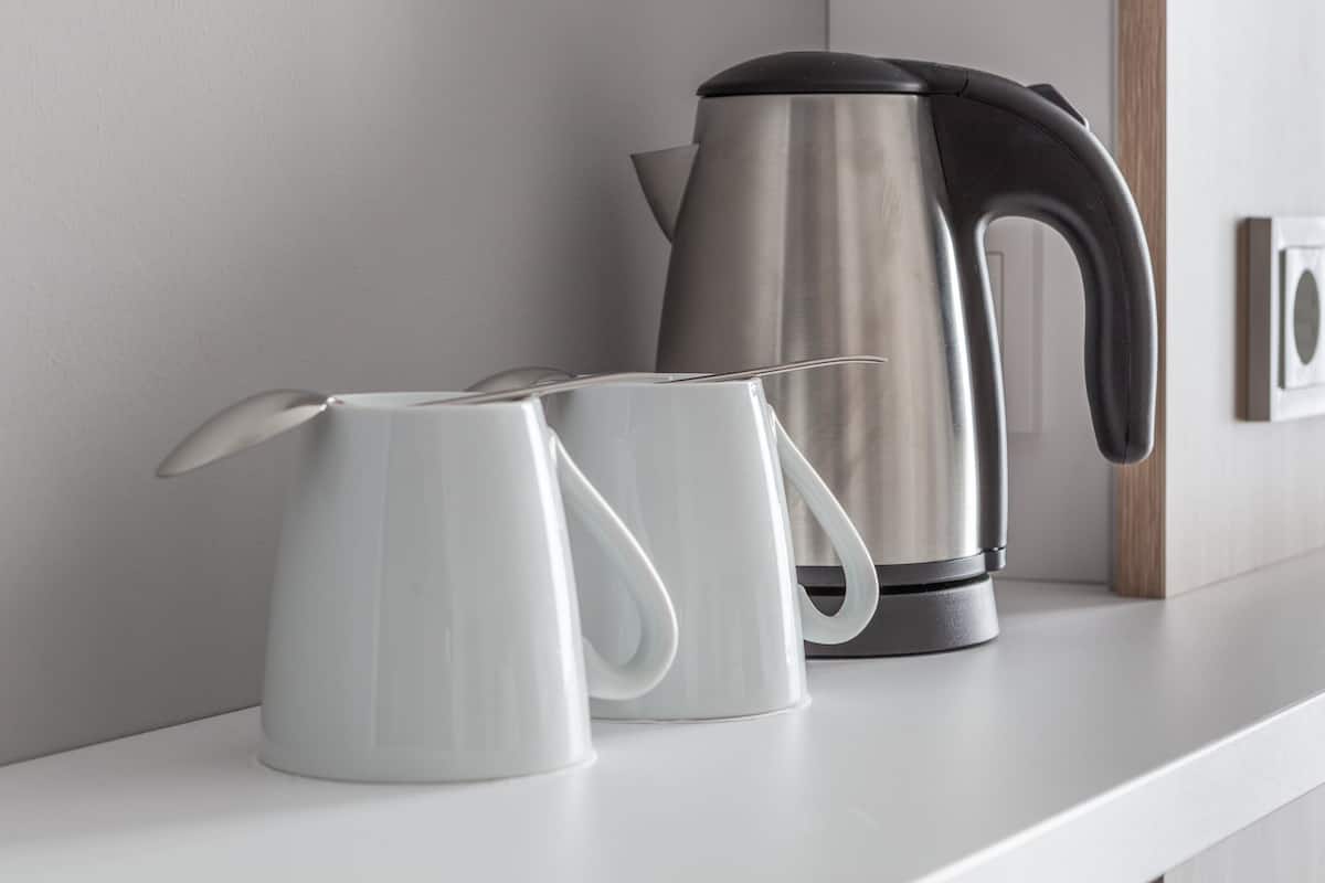 A white shelf with a stainless steel kettle and two white mugs.