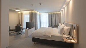 Una habitación de hotel moderna con una cama grande, un escritorio con televisor de pantalla plana, una silla y una mesita con teléfono y documentos.