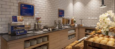 Un'area colazione a buffet con angolo dedicato ai pancake, distributori di frutta e cereali, e un'ampia scelta di pane e pasticceria.