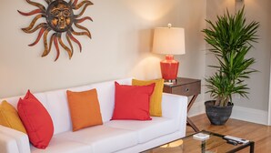 En hvid sofa med røde og orange puder, et glas sofabord, en rød lampe, en potteplante og en soldekoration på væggen.