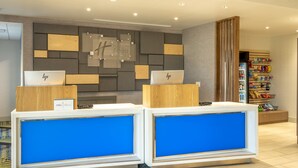 A hotel reception area with two HP laptops on desks, a Holiday Inn Express sign, and a vending machine with snacks. - Holiday Inn Express & Suites Watertown by IHG (Watertown)