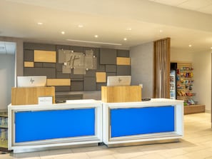 A hotel reception area with two HP laptops on desks, a Holiday Inn Express sign, and a vending machine with snacks. - Holiday Inn Express & Suites Watertown by IHG (Watertown)
