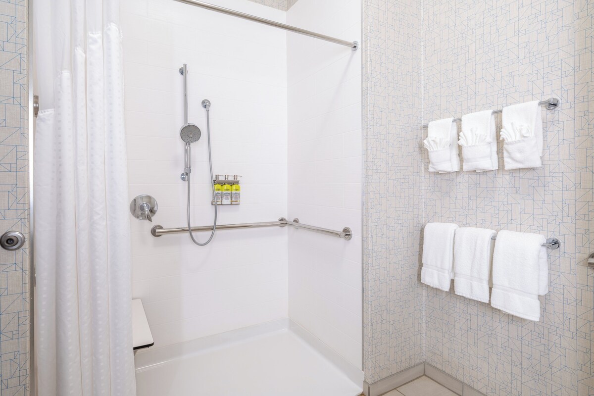 A walk-in shower with a handheld showerhead, a handheld showerhead, and a grab bar. There are three white towels hanging on a towel rack.