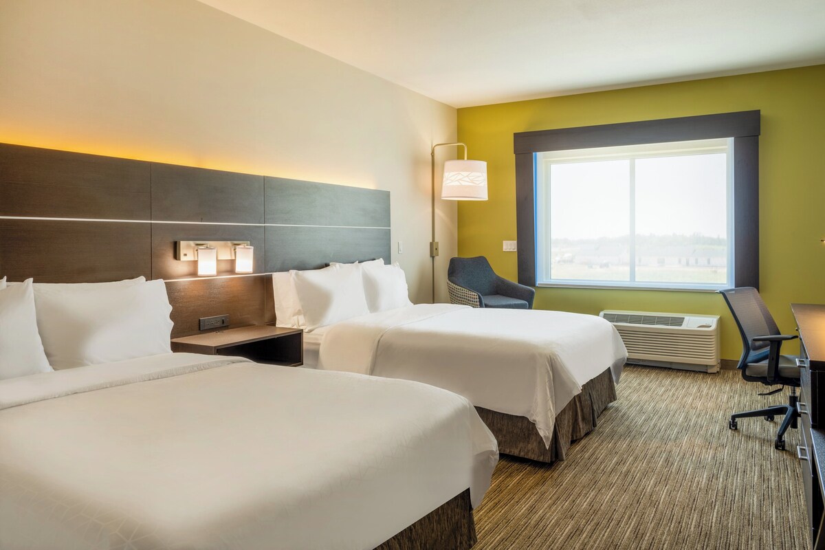 A hotel room with two beds, a desk, a chair, a window with a view, and a wall-mounted lamp.