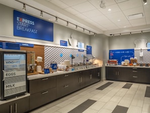 A breakfast area with a yogurt machine, cereal, and hot food options. - Holiday Inn Express & Suites Watertown by IHG (Watertown)