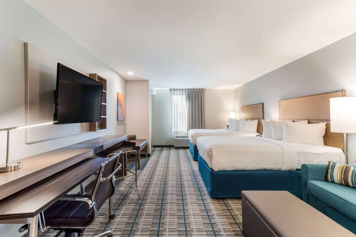 A hotel room with a large bed, a desk with a chair, a TV mounted on the wall, and a window with curtains.