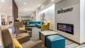 Un vestíbulo de hotel moderno con un letrero de 'Welcome', asientos cómodos, una chimenea y una cocina al fondo.