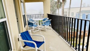 A balcony with white tables and blue chairs, a black railing, and a view of palm trees and buildings in the background.