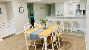 A dining area with a wooden table and white chairs, a kitchen with white cabinets, and a clock on the wall.