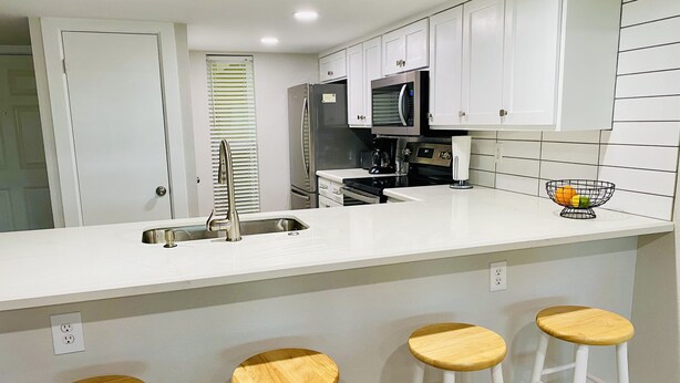 A kitchen with white cabinets, a stainless steel refrigerator, a microwave, a stove, a sink, and four wooden stools.