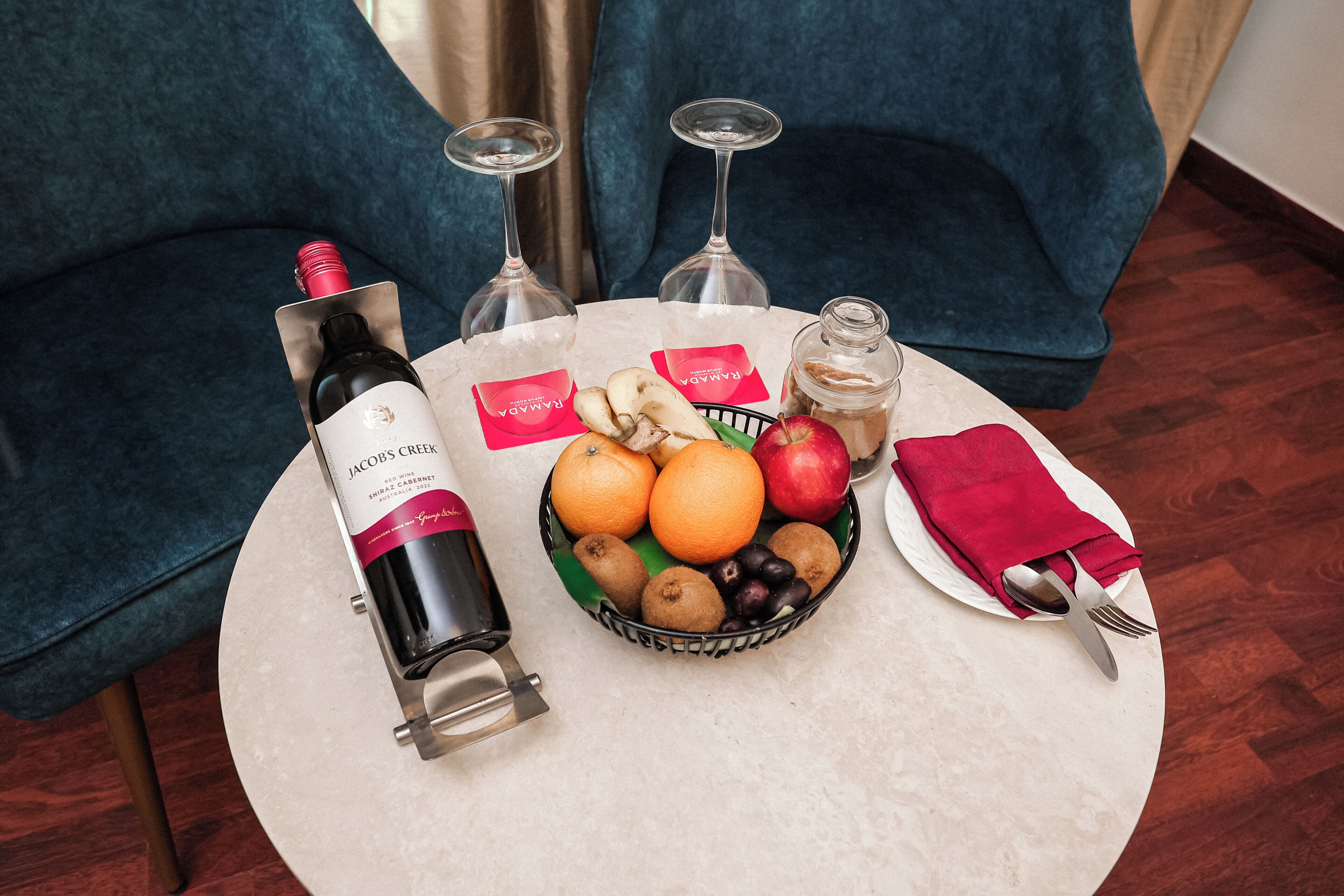 A table with a bottle of wine, glasses, a bowl of fruit, and a jar on a round table.