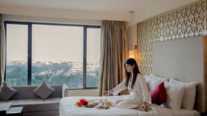 Habitación de hotel con cama, sofá, mesa de desayuno y un amplio ventanal con vista a la ciudad.