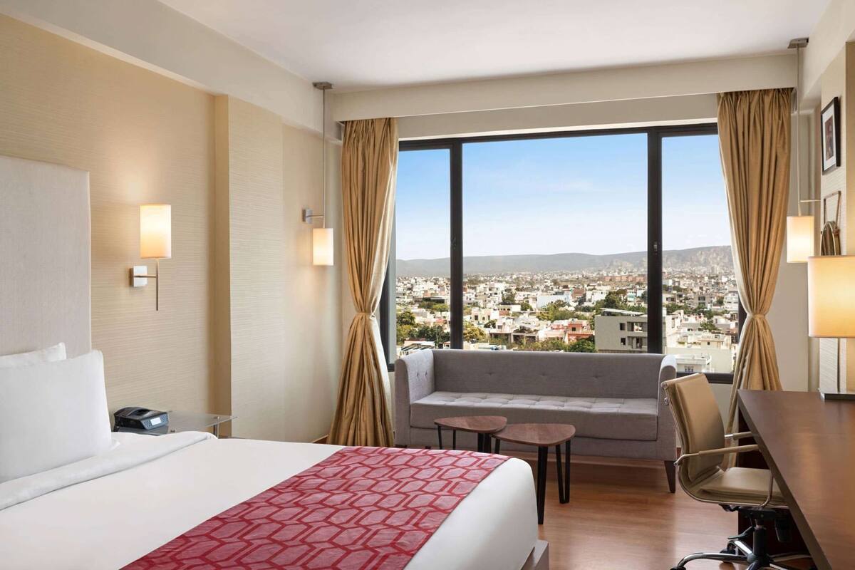 A hotel room with a bed, a sofa, a desk, and a view of a cityscape.