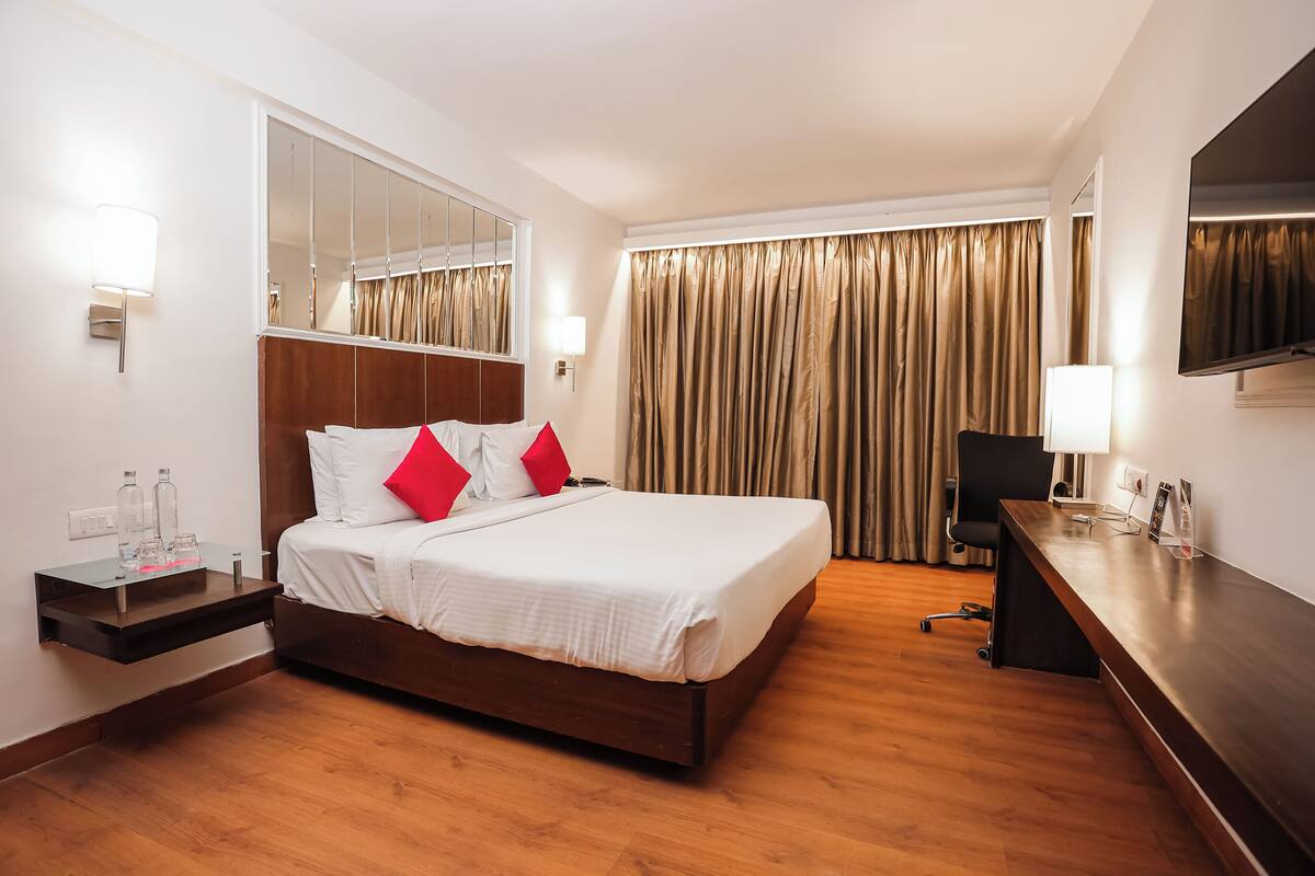 A hotel room with a bed, bedside table, lamp, mirror, and a desk with a chair.