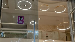 Una mujer con vestido verde camina con una maleta en un moderno vestíbulo de hotel con paredes de vidrio, luces circulares en el techo y un cartel que dice 'Use máscara facial'.