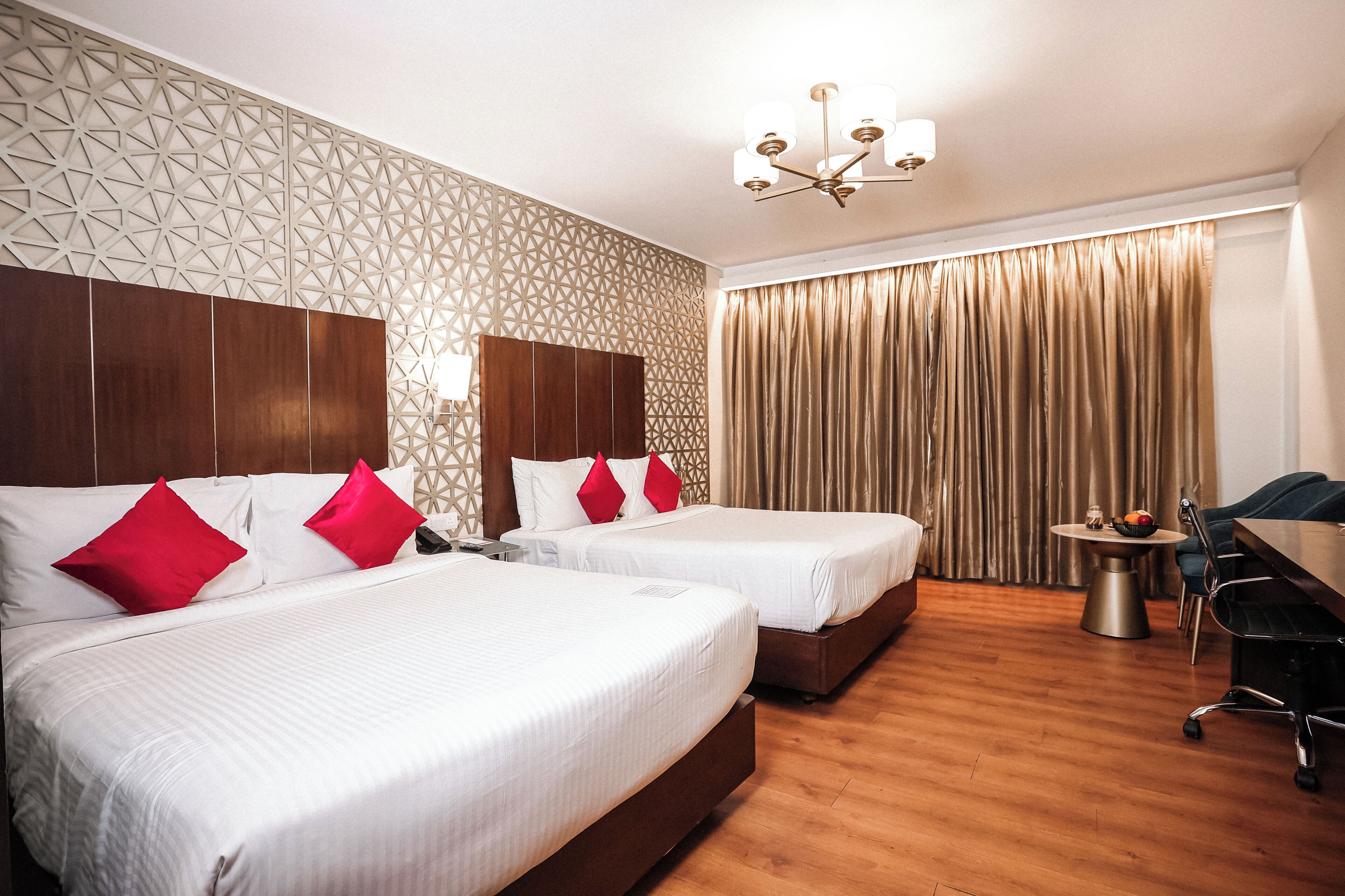 A hotel room with two beds, a desk, a chair, a chandelier, and a window with curtains.