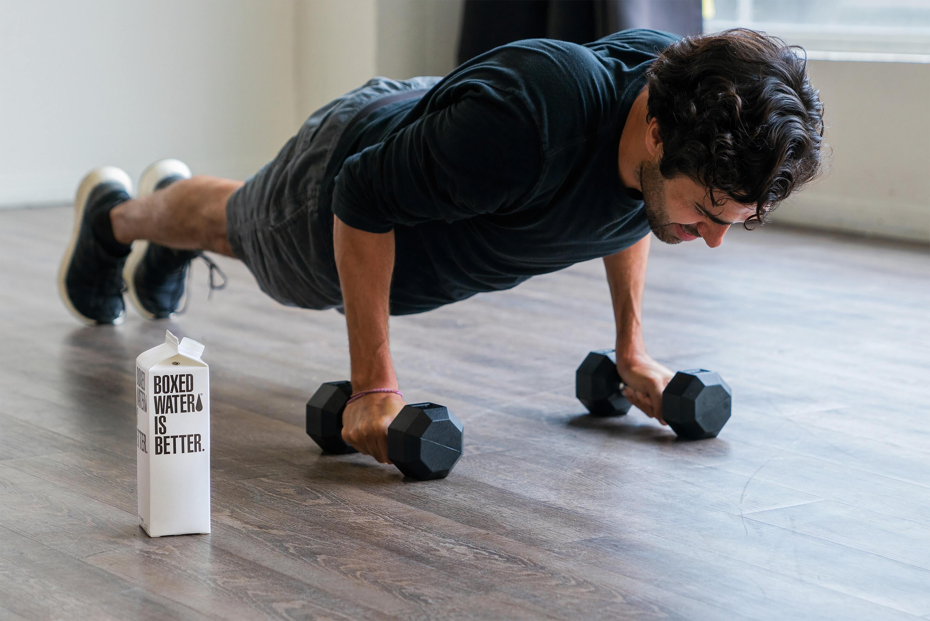 A person doing push-ups with dumbbells on a wooden floor, and a carton of boxed water nearby.