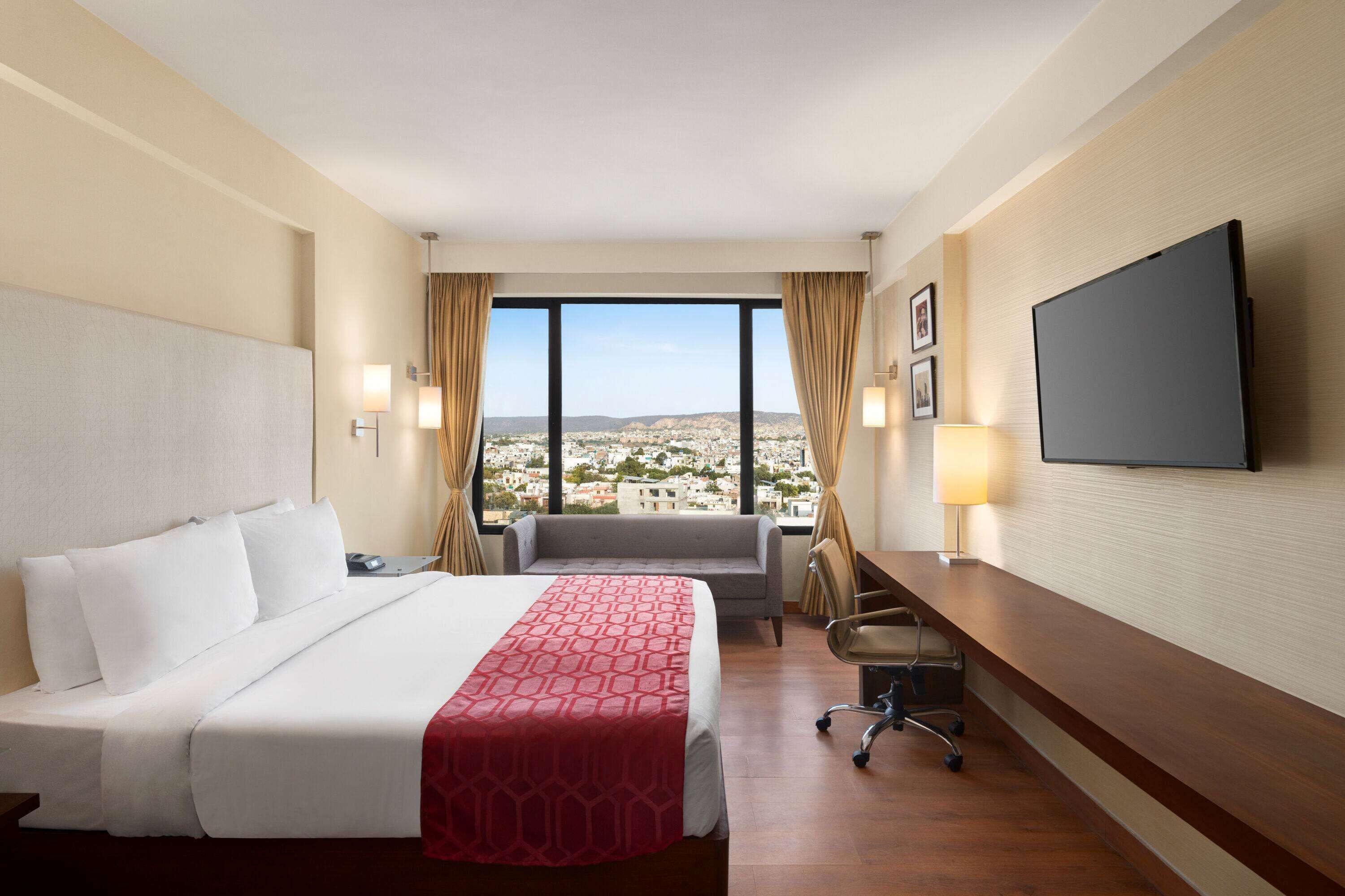 Habitación de hotel con cama, escritorio, silla, televisor y vistas a la ciudad.