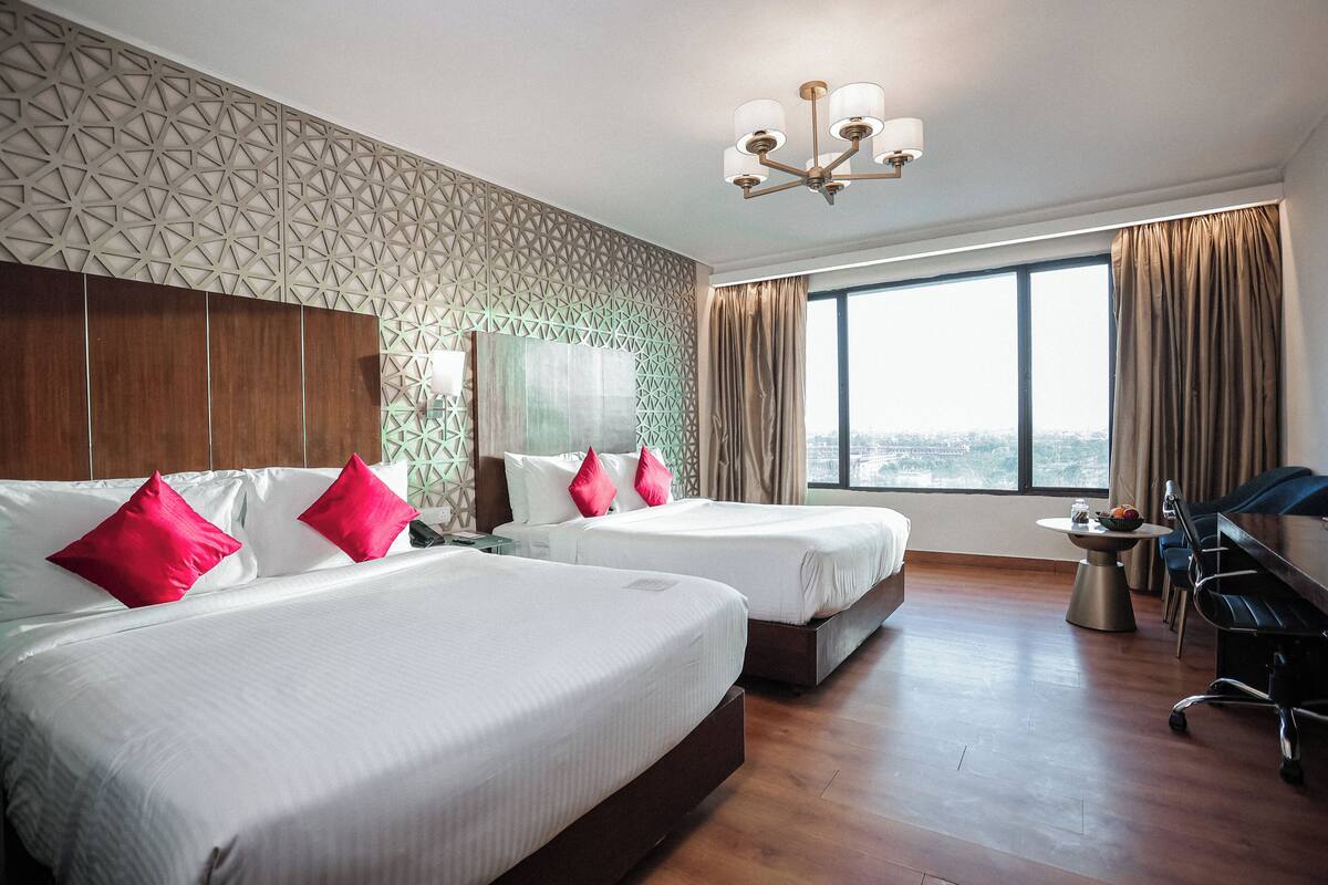 A hotel room with two beds, a desk, a chair, a window with a city view, and a geometric wall pattern.