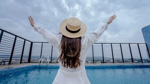 Una persona de pie junto a una piscina, con los brazos levantados, vestida con un conjunto blanco y un sombrero de paja.