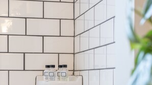 Et badeværelse med hvide fliser, en brusehoved og en hylde med toiletartikler.