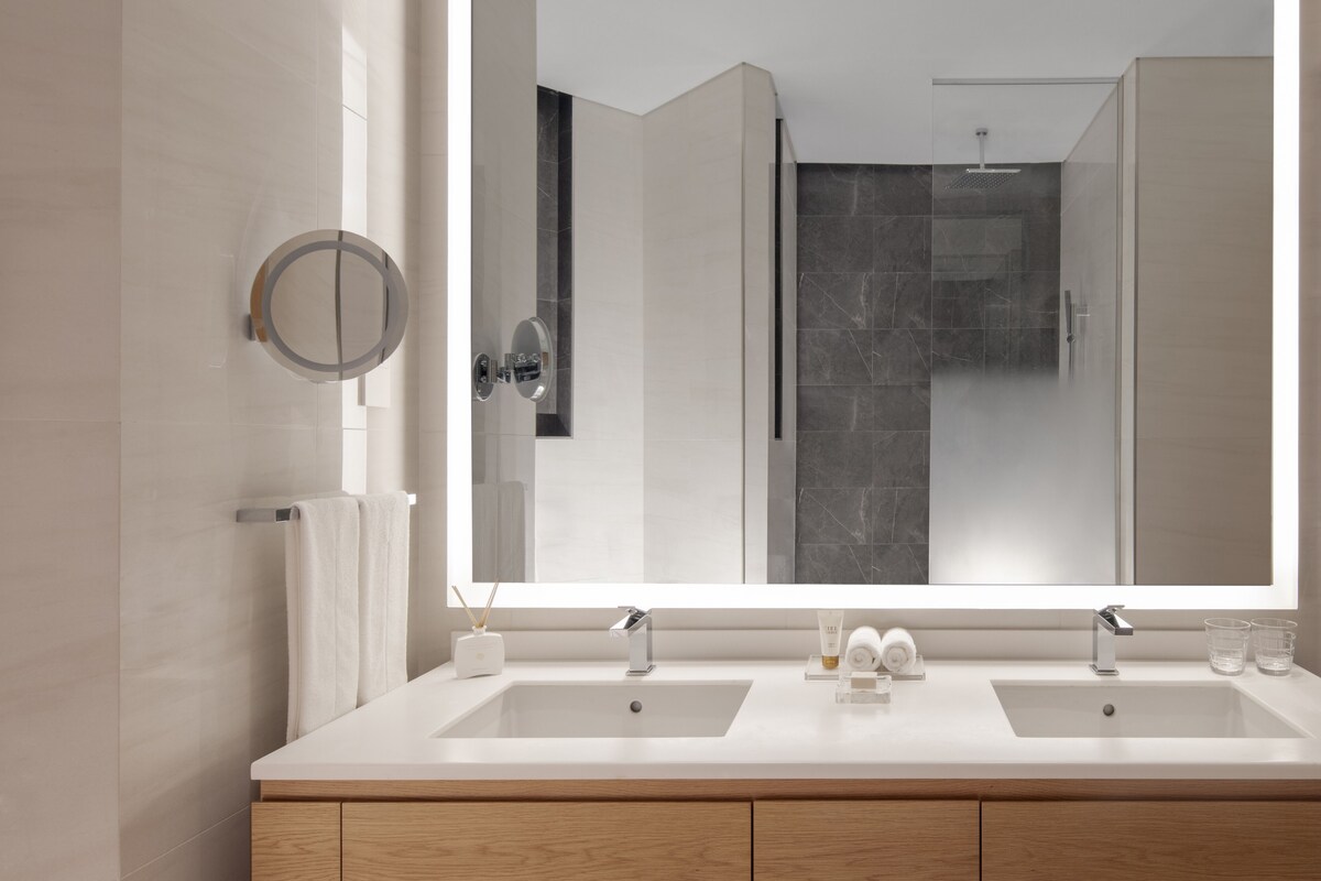 A modern bathroom with a large mirror, two sinks, and a shower visible in the reflection.