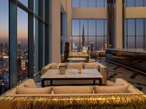 Une chambre d’hôtel située dans un immeuble de grande hauteur, offrant une vue sur le panorama urbain, avec un coin repas comprenant une table et des chaises, et un espace détente avec des canapés et un tapis à motifs.