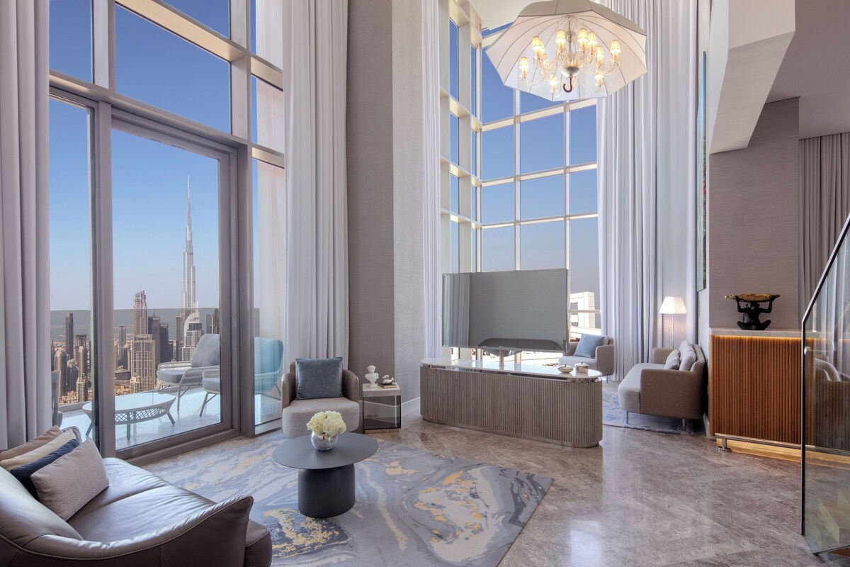 A spacious living room with a large window offering a city view, a modern chandelier, and a comfortable seating area.
