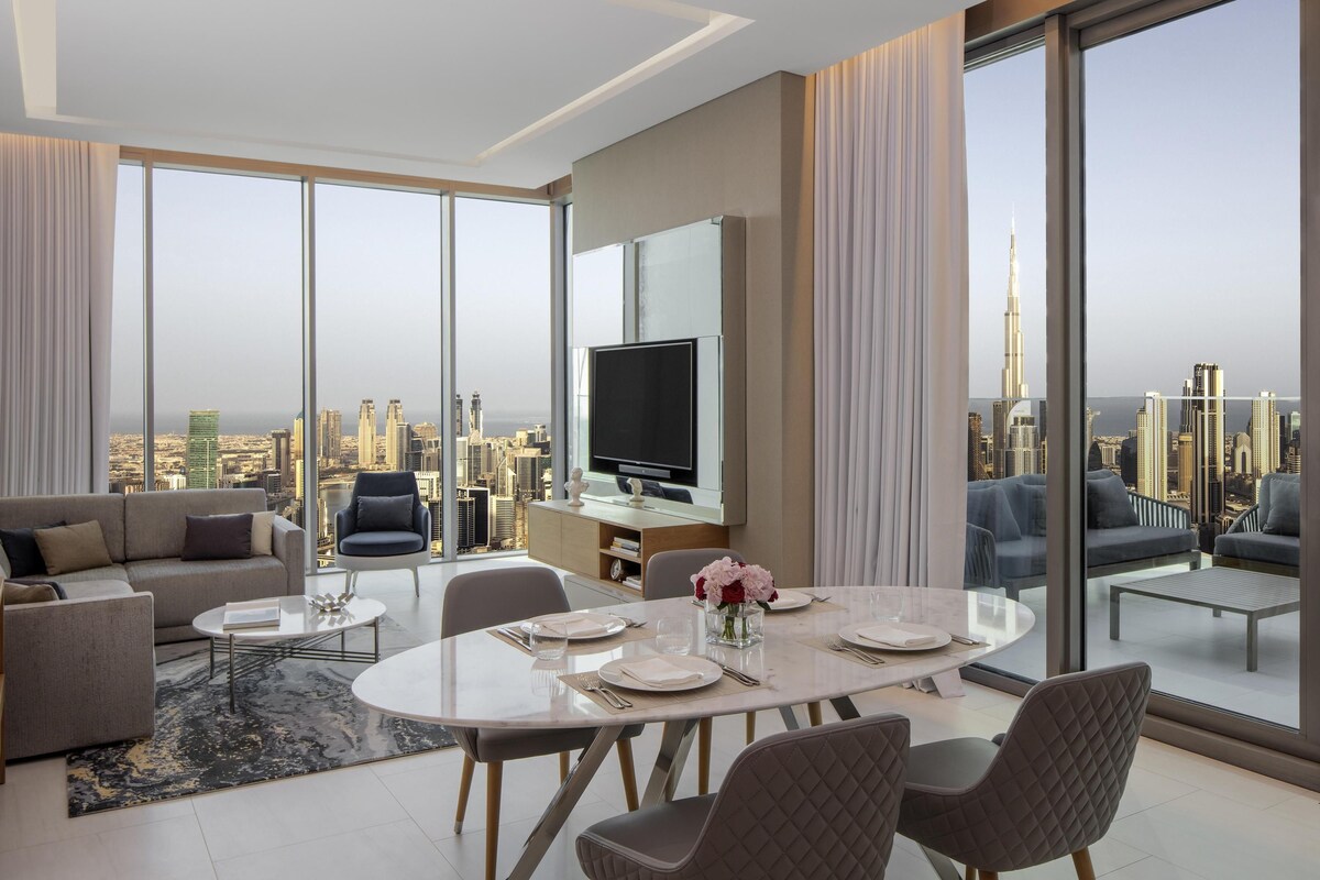 A modern living room with a dining area, large windows offering a city view, a flat-screen TV, and a sofa.