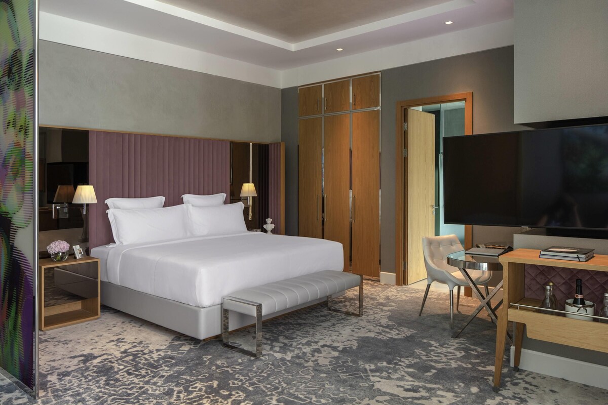 A modern hotel room with a large bed, bedside tables, a desk with a chair, a television, and a wardrobe.