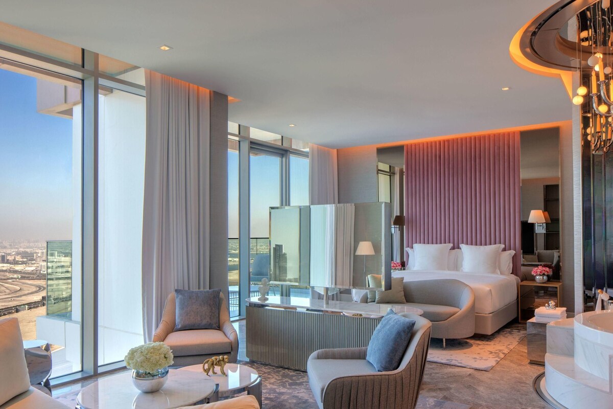 A modern hotel room with a large bed, a sitting area with a coffee table, and a view of the cityscape.
