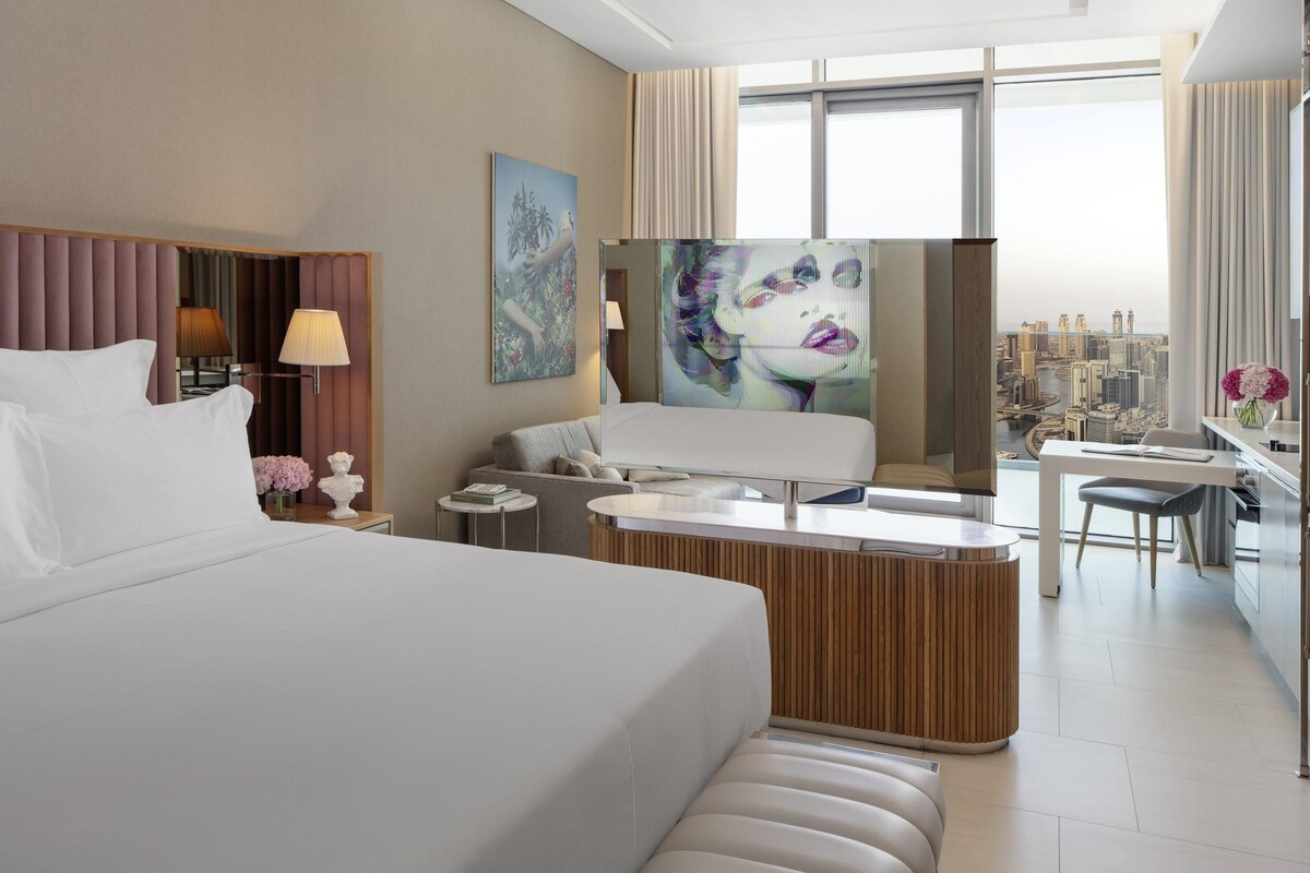 A modern hotel room with a large bed, a desk, and a view of the city.