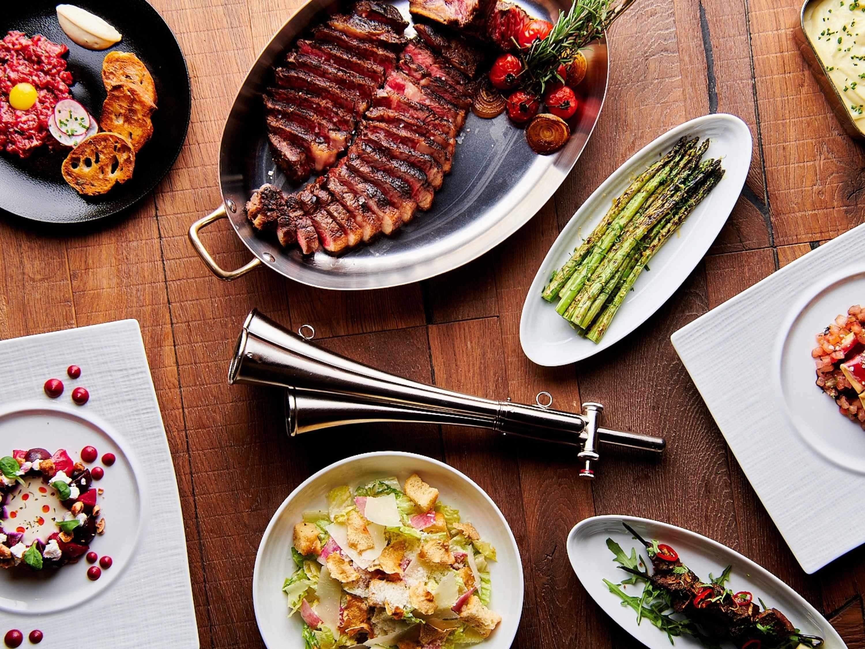 A variety of dishes including steak, asparagus, and a salad, arranged on a wooden table.