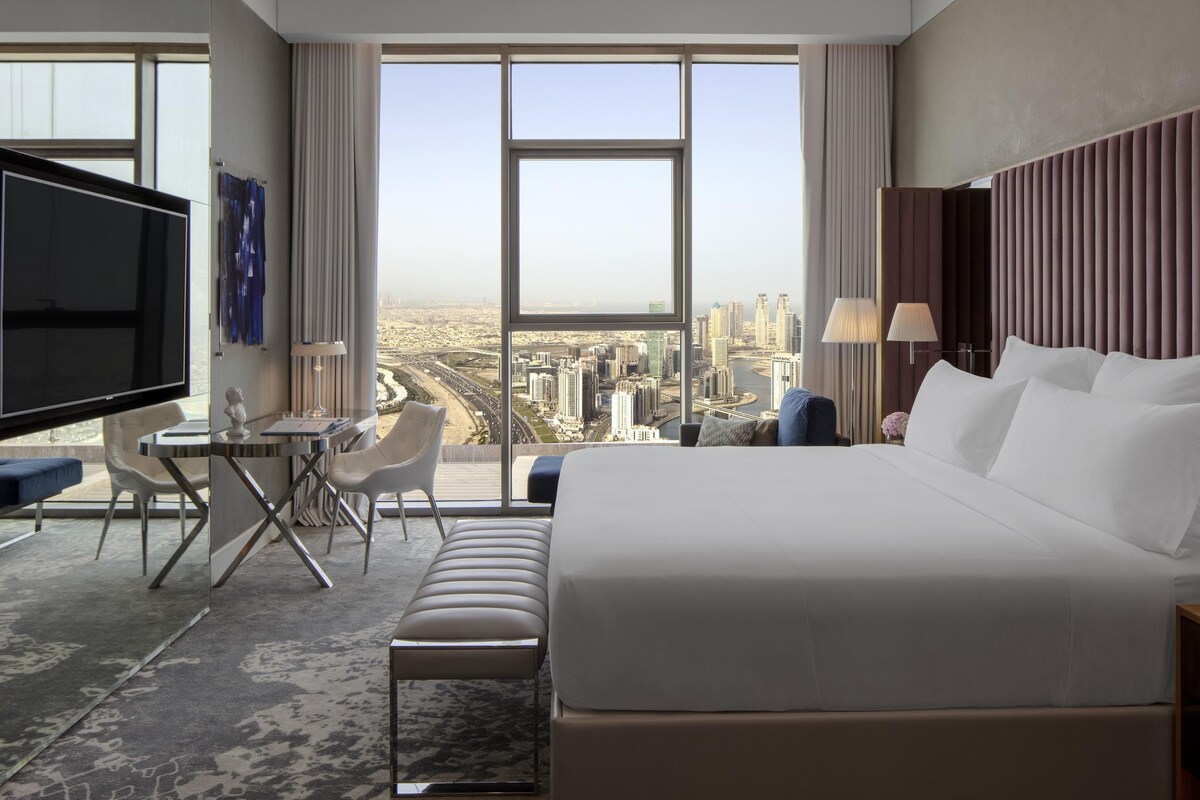 A modern hotel room with a large bed, a desk, a TV, and a city view.