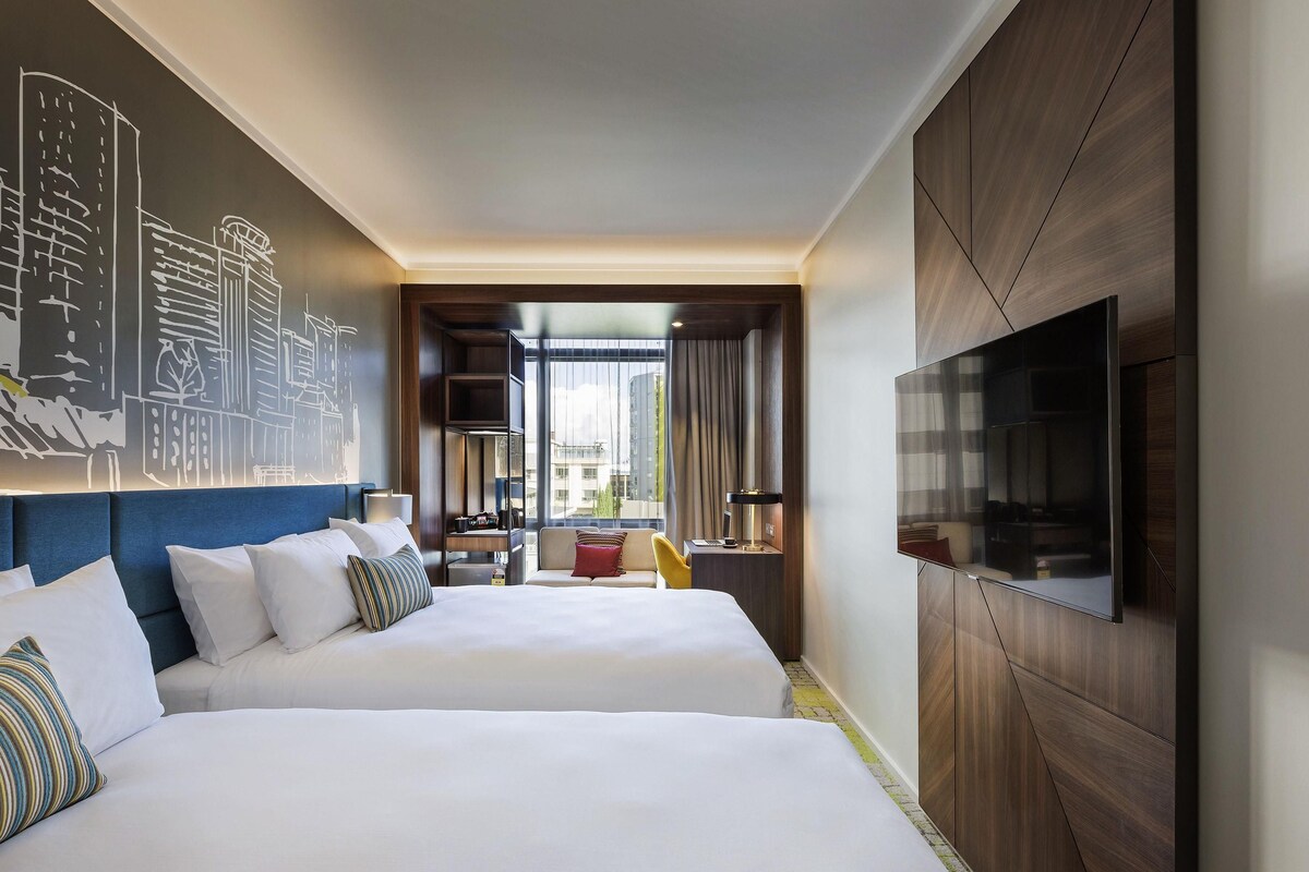 A hotel room with two beds, a TV, a bookshelf, and a cityscape mural.