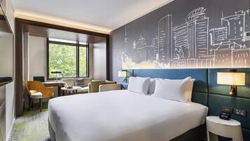 Habitación de hotel con una cama grande, un escritorio con una silla, una ventana con vistas a árboles y un mural de la ciudad en la pared.