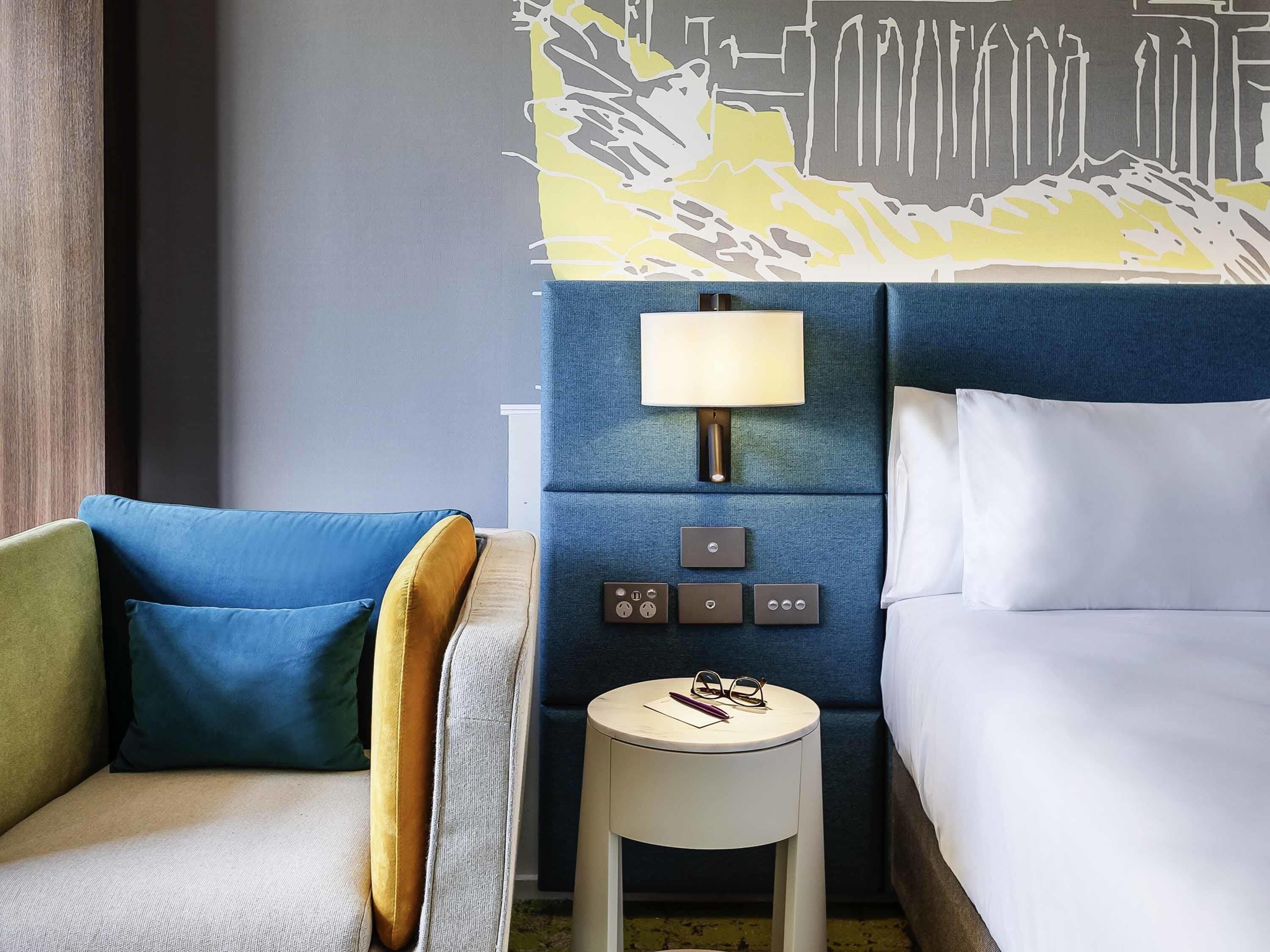 Une chambre d’hôtel avec un lit, une table de chevet avec une lampe, une chaise et un mur orné d’une fresque représentant un paysage urbain.