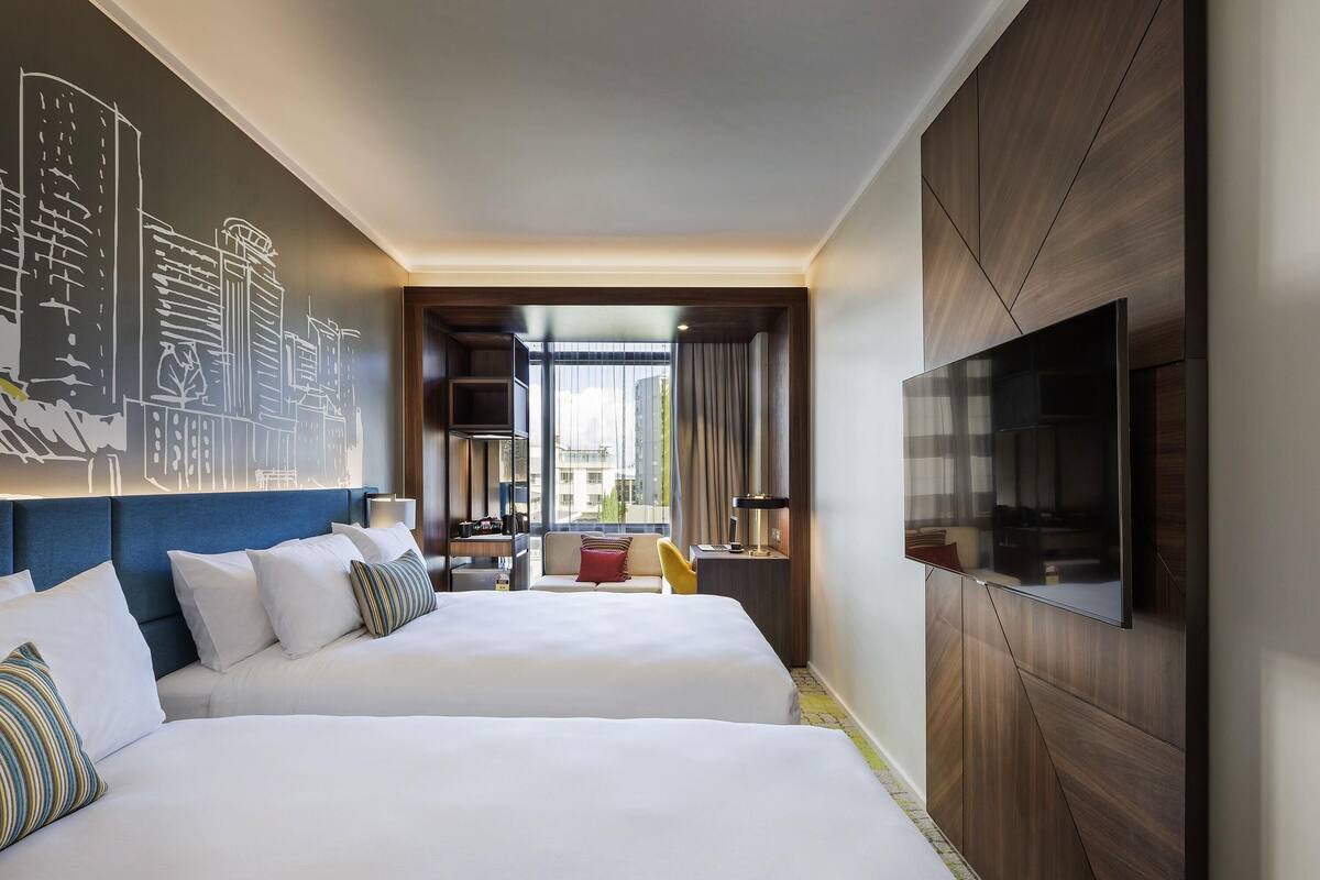 A hotel room with two beds, a TV, a bookshelf, and a cityscape mural.
