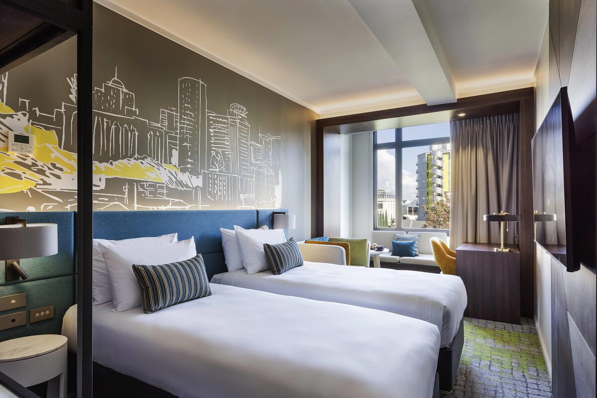 A hotel room with two beds, a cityscape mural, a desk with a lamp, and a view of buildings through the window.
