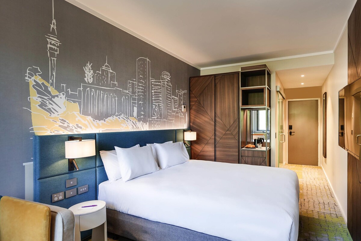 A hotel room with a large bed, a cityscape mural on the wall, a desk with a lamp, and a wardrobe.